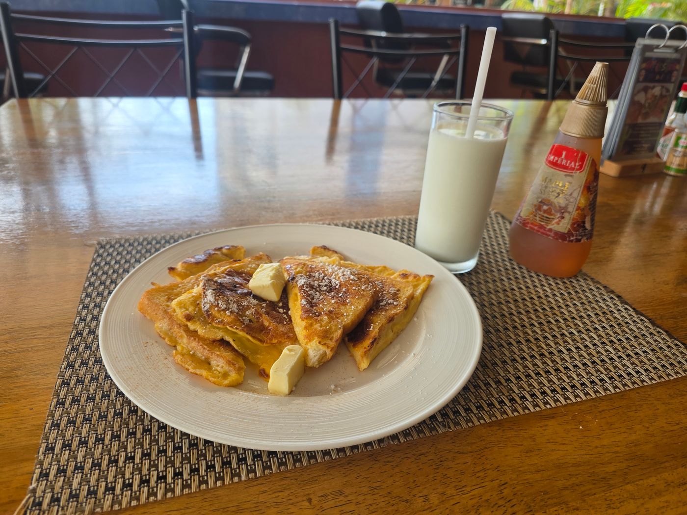 French toast with butter and powdered sugar, a glass of milk, and maple syrup on a wooden bar table in Pattaya, photographed by Danny Flight.